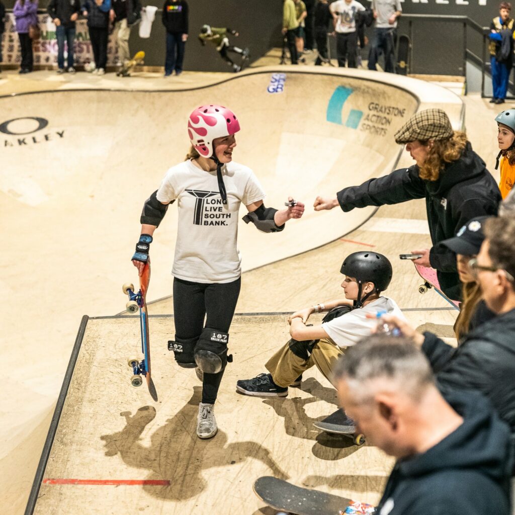 GB's Skateboard Pipeline Project, Young skateboarder holding a skateboard with others on the side of a skate bowl